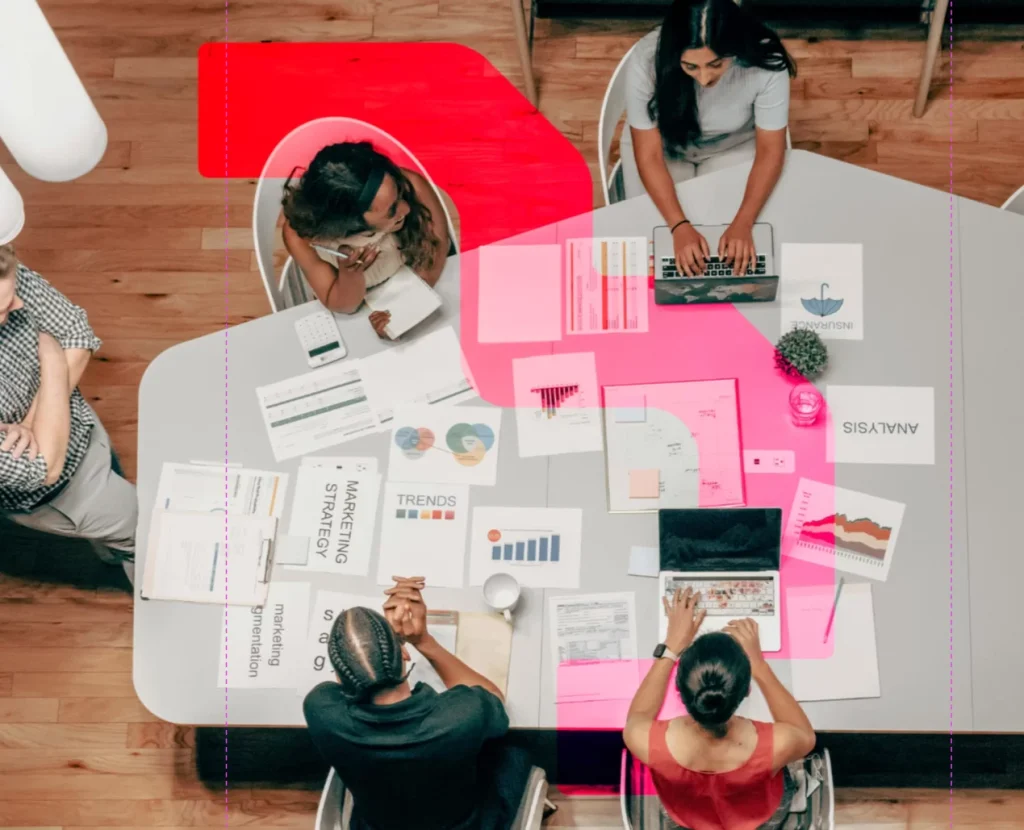 Overhead view of a digital marketing team collaborating around a meeting table with laptops, charts, reports and strategy documents. Team members analyse marketing performance, campaign data and business insights during a planning session in a modern office.